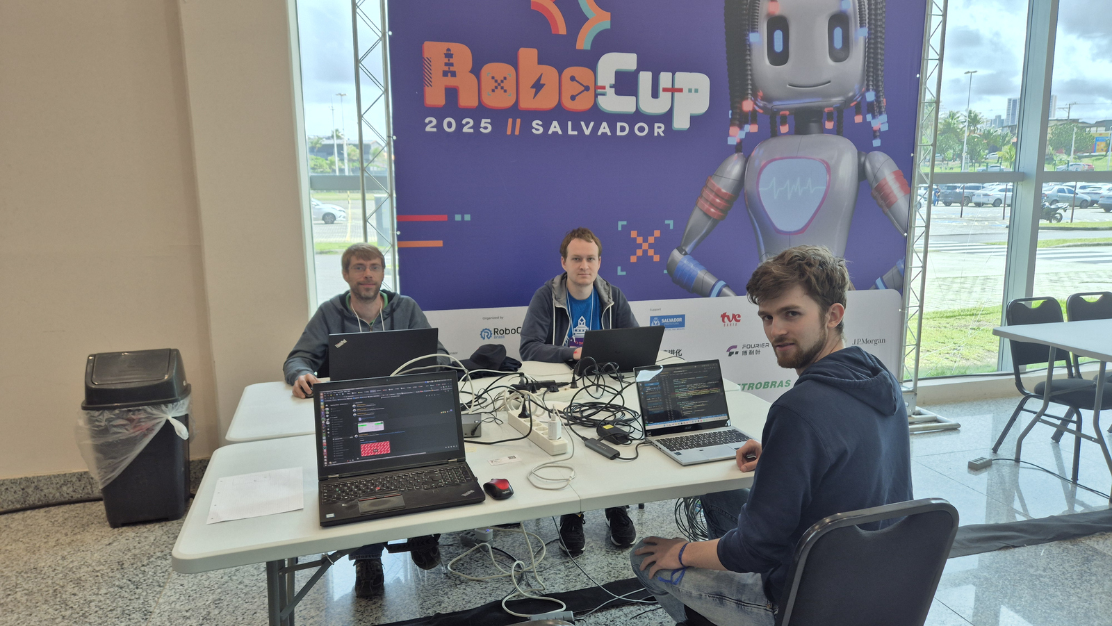 Drei junge Männer sitzen um einen Tisch mit Laptops im Hintergrund ist ein RoboCup-BAnner zu sehen