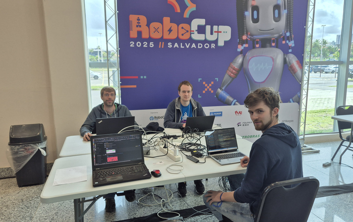 Drei junge Männer sitzen um einen Tisch mit Laptops im Hintergrund ist ein RoboCup-BAnner zu sehen