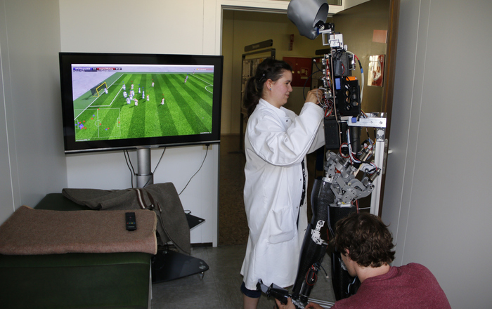 Zwei Studierende arbeiten im Sanitätsraum der Hochschule Offenburg daran, Sweaty wieder fit zu machen während dieser auf einen Bildschirm schaut auf dem ein Spiel des Teams Magma läuft.