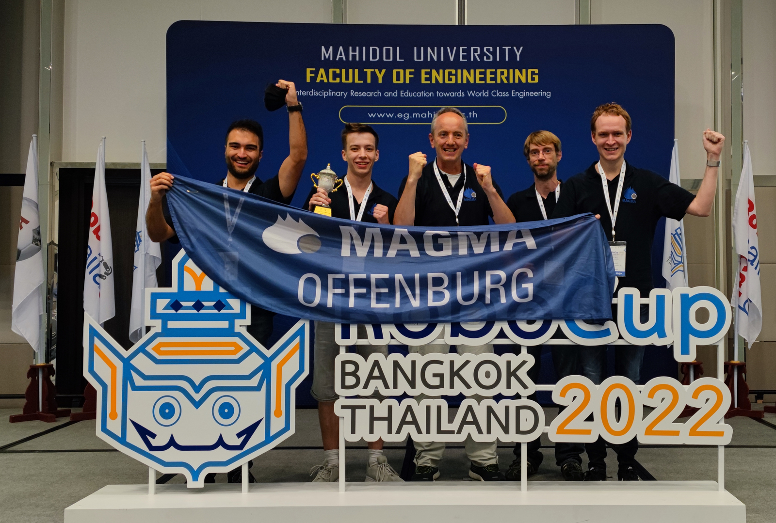 Das Team Magma jubelt mit dem Siegerpokal und einer Teamflagge hinter einem Logo des RoboCups Bangkok