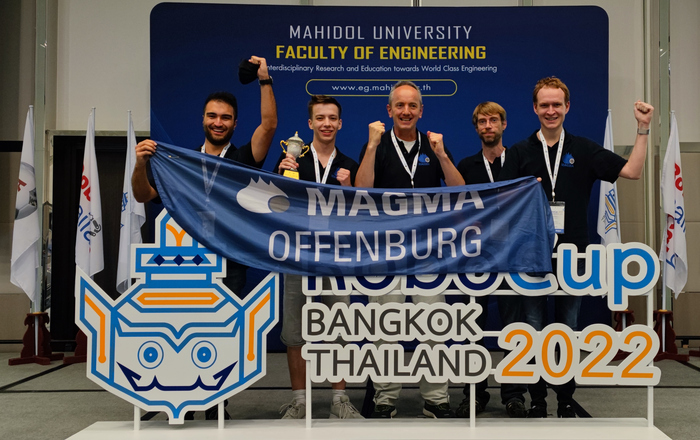 Das Team Magma jubelt mit dem Siegerpokal und einer Teamflagge hinter einem Logo des RoboCups Bangkok