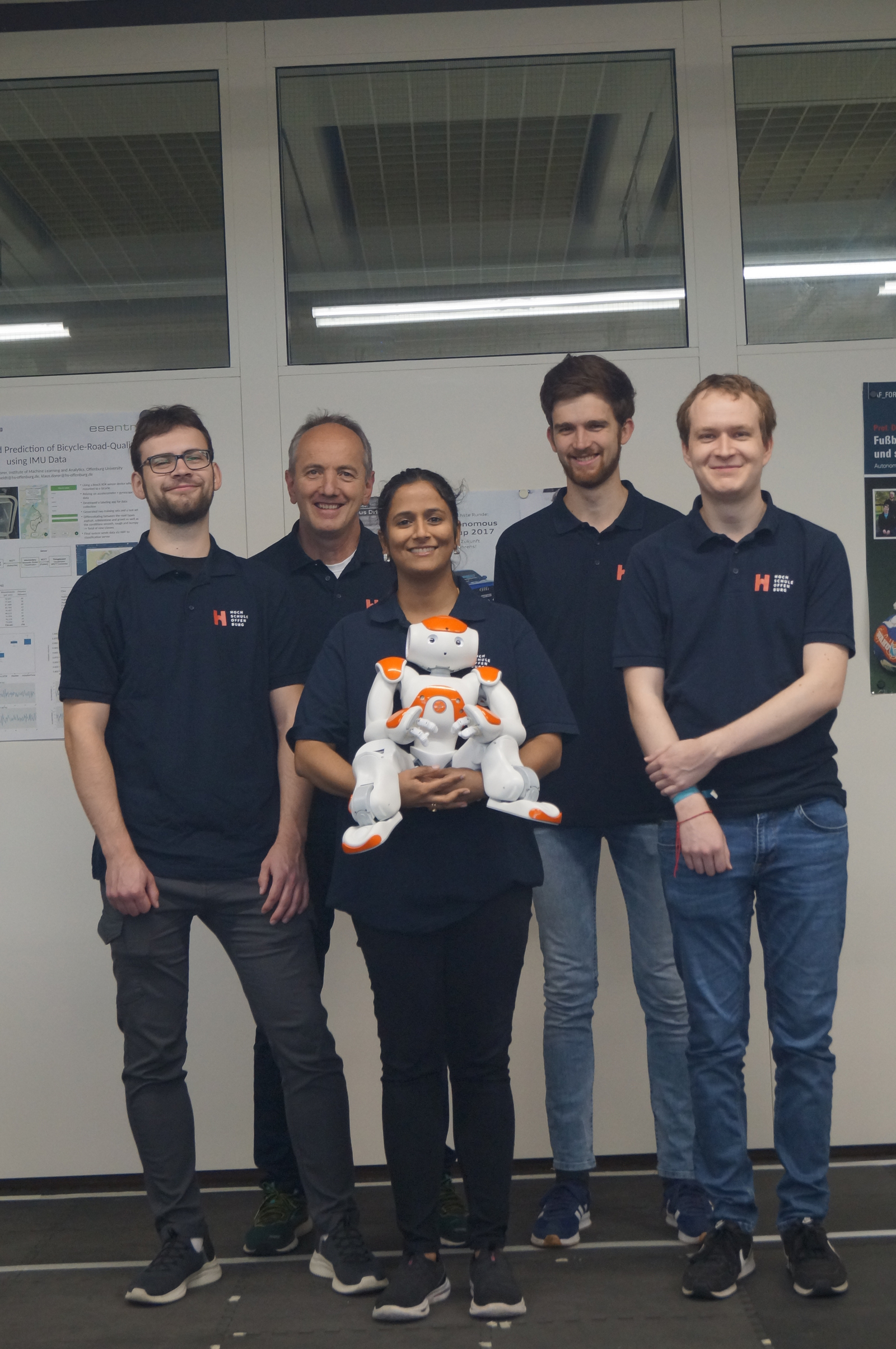 Vier junge Männer und eine Frau stehen in einer Reihe, die Frau in der Mitte hält einen Nao-Roboter